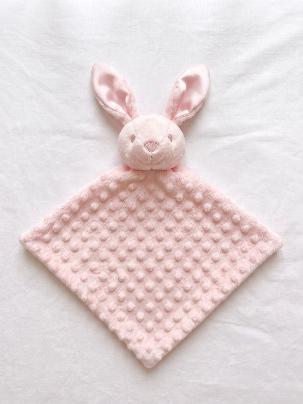 A light pink bunny security blanket with a textured dotty fabric body and a plush bunny head, laid flat on a white fabric background.