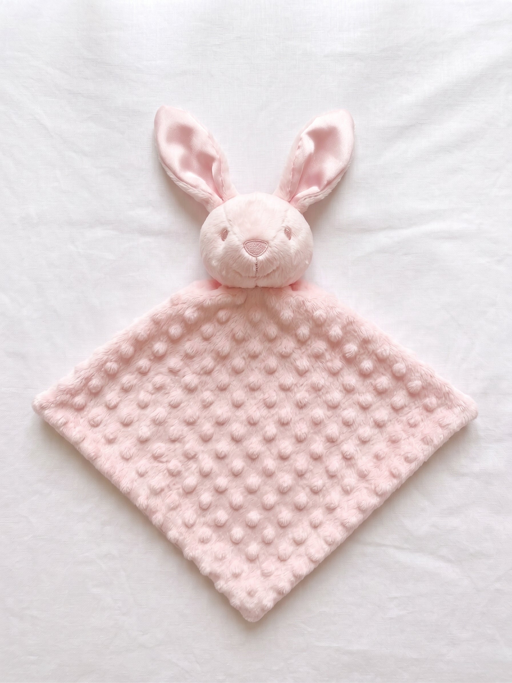 A light pink bunny security blanket with a textured dotty fabric body and a plush bunny head, laid flat on a white fabric background.