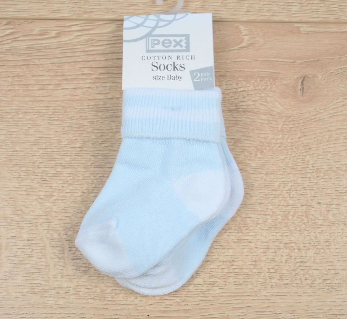 A 2-pack of light blue cotton-rich baby socks with white heels and toes, hanging from a white PEX brand paper header card on a light wood background.