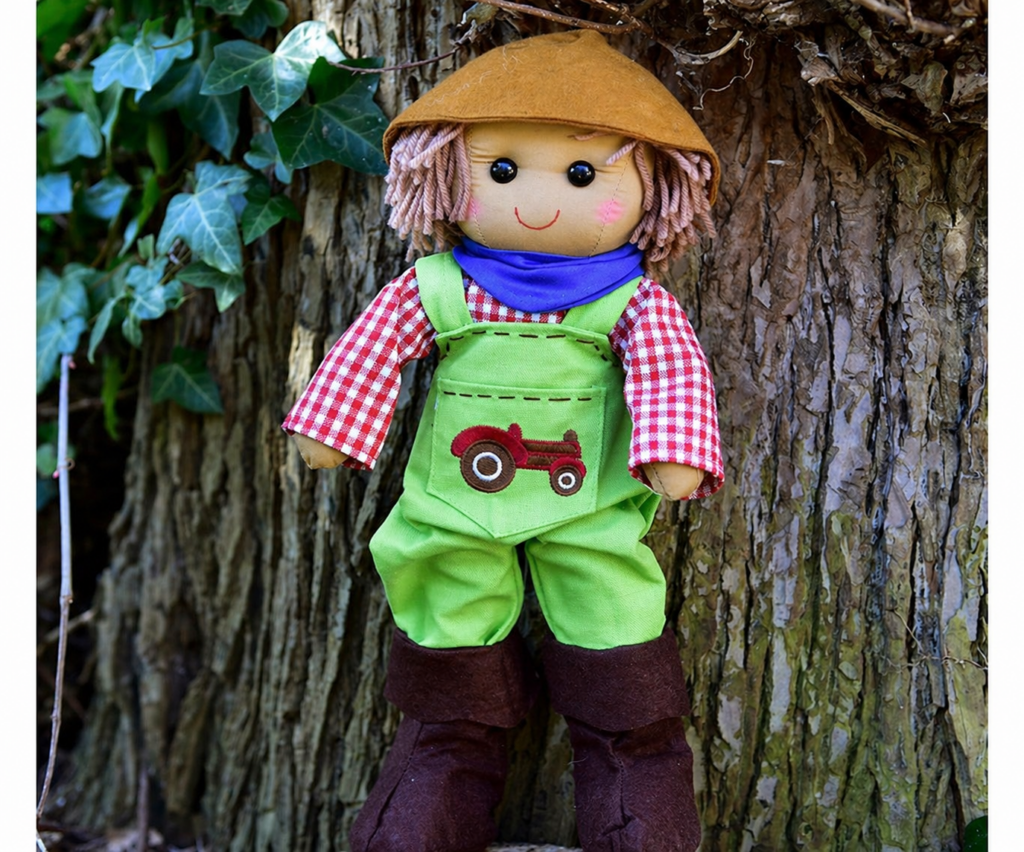 Farmer rag doll in green dungarees with embroidered tractor, red gingham shirt, blue neckerchief and tan hat, leaning against a tree