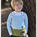 Young boy wearing baby blue tractor knitted jumper with farmyard scene, smiling against a tree