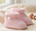 A pair of light pink hand-knit baby booties with a small matching pink bow on one bootie, displayed on a soft cream knit blanket.