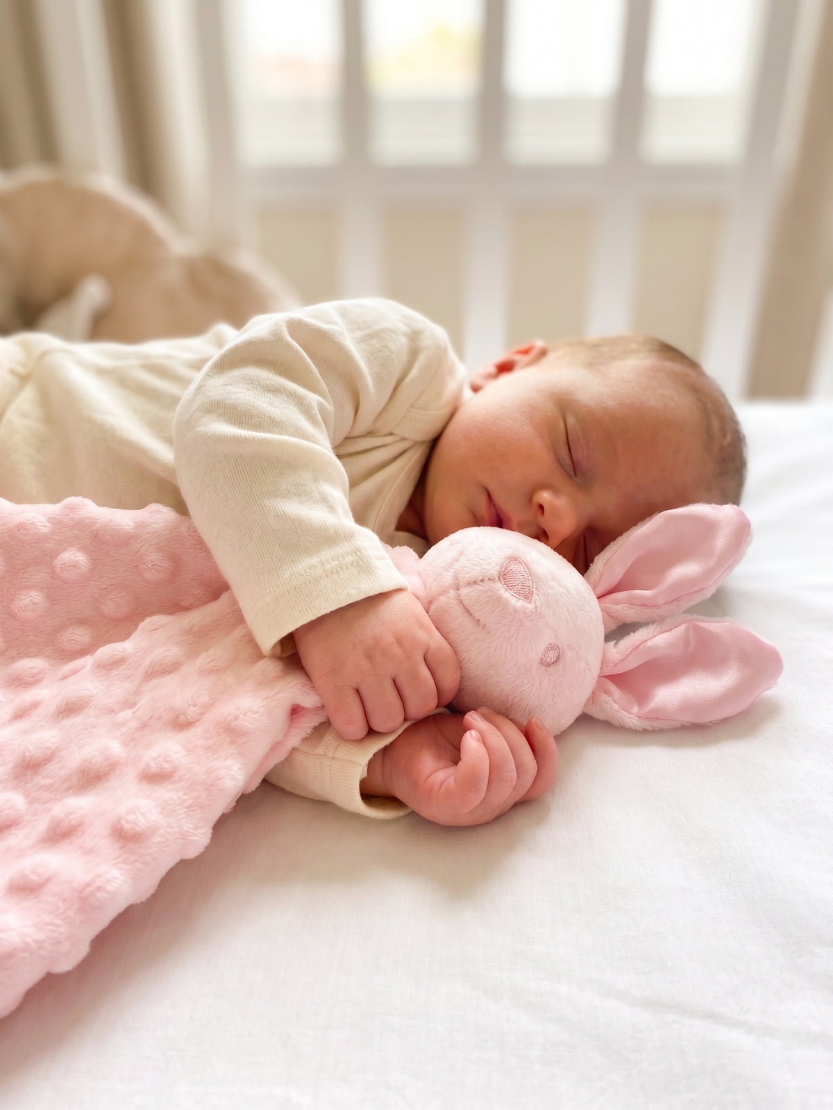 A sleeping newborn baby in a cream onesie rests on a white bed, holding a pink bunny-shaped plush toy with a pink dotted blanket.