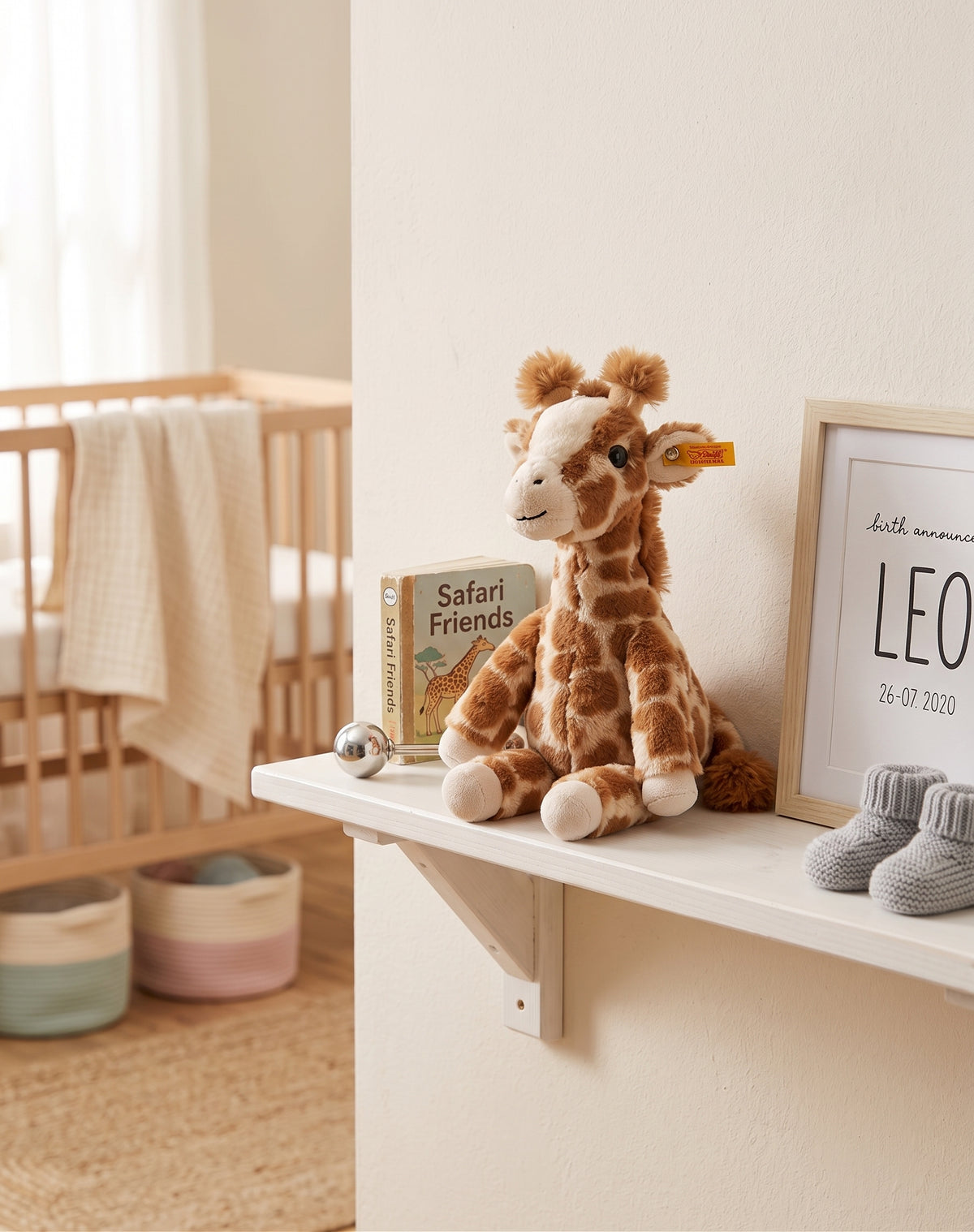 A plush giraffe toy sits on a white shelf in a softly styled nursery. A wooden crib with a cream blanket is visible in the background, along with a small book, a silver ball, a framed print, and a pair of gray baby booties.